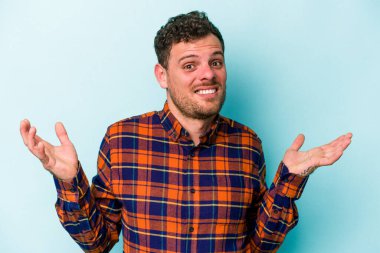 Kafkasyalı genç adam mavi arka planda izole edilmiş sorgulama jestinde şüphe ediyor ve omuz silkiyor..