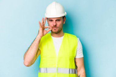 Young laborer caucasian man isolated on blue background with fingers on lips keeping a secret.