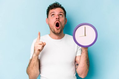 Kafkasyalı genç bir adam mavi arka planda izole edilmiş bir saat tutuyor ve ağzı açık bir şekilde üst tarafı işaret ediyor..