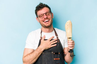 Kafkasyalı genç kunduracı, mavi arka planda izole edilmiş. Yüksek sesle gülüyor, elini göğsüne koyuyor..