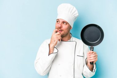 Kafkasyalı genç aşçı elinde uçan tavayla mavi arka planda izole edilmiş bir şekilde duruyor. Bir şeyin fotokopi odasına baktığını düşünüyor..