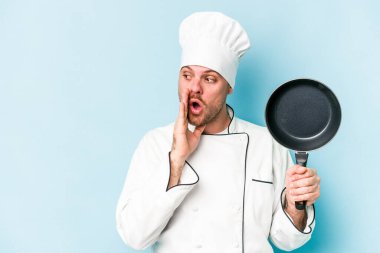 Kafkasyalı genç aşçı elinde uçan bir tavayla mavi arka planda izole edilmiş bir şekilde gizli bir sıcak fren haberi veriyor ve yana bakıyor.