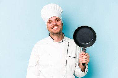 Kafkasyalı genç aşçı elinde uçan tavayla mavi arka planda izole edilmiş mutlu, güler yüzlü ve neşeli.
