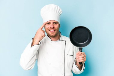 Kafkasyalı genç aşçı elinde uçan tavayla mavi arka planda izole edilmiş işaret parmağıyla hayal kırıklığı jesti yapıyor..