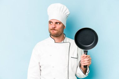Kafkasyalı genç aşçı elinde uçan tavayla mavi arka planda izole edilmiş, kafası karışmış, şüpheli ve güvensiz hissediyor..