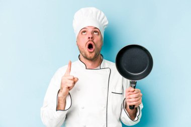 Kafkasyalı genç aşçı elinde uçan tavayla mavi arka planda izole edilmiş ağzı açık bir şekilde yukarı işaret ediyor..