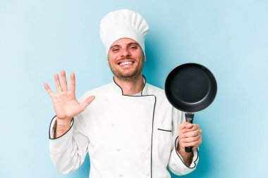 Kafkasyalı genç aşçı elinde uçan tavayla mavi arka planda izole edilmiş gülümseyen beş numarayı parmaklarıyla gösteriyor..