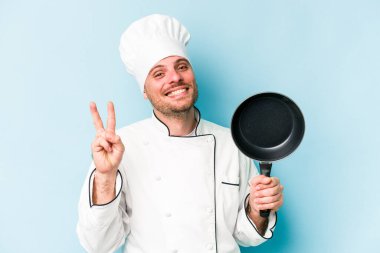 Kafkasyalı genç aşçı elinde uçan tavayla mavi arka planda izole edilmiş neşeli ve tasasız parmaklarıyla barış sembolü gösteriyor..