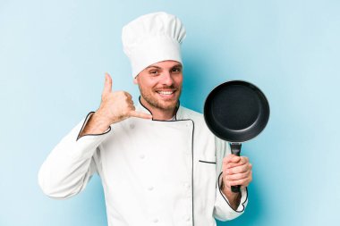 Kafkasyalı genç aşçı elinde uçan tavayla mavi arka planda izole edilmiş bir şekilde parmaklarıyla bir cep telefonu hareketi gösteriyor..