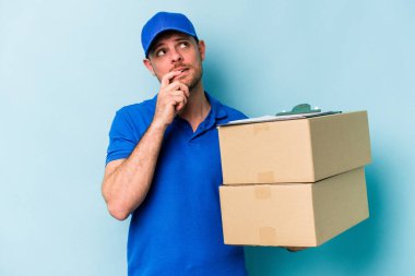 Kafkasyalı genç teslimatçı mavi arka planda izole edilmiş rahat bir şekilde fotokopi odasına bakan bir şey düşünüyor..