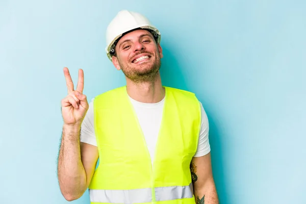 Young laborer caucasian man isolated on blue background joyful and carefree showing a peace symbol with fingers.
