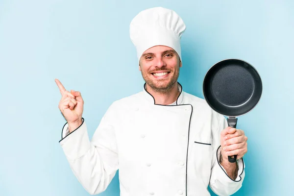 年轻的高加索厨师牵着被蓝色背景隔开的飞行锅 微笑着指着旁边 在空旷的空间里展示着一些东西 — 图库照片