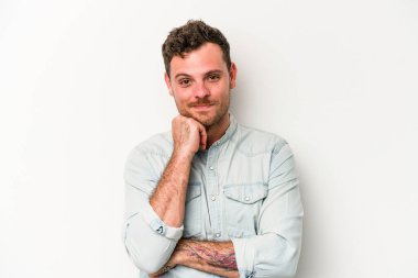 Beyaz arka planda izole olmuş genç beyaz bir adam gülümsüyor ve kendinden emin, çenesine elleriyle dokunuyor..