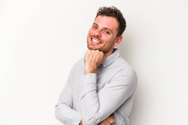 Beyaz arka planda izole olmuş genç beyaz bir adam gülümsüyor ve kendinden emin, çenesine elleriyle dokunuyor..