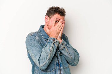 Beyaz arka planda izole edilmiş genç beyaz bir adam parmaklarının arasından korkmuş ve gergin bir şekilde göz kırpıyor..