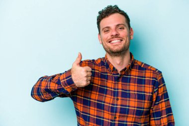 Mavi arka planda izole edilmiş genç beyaz adam gülümsüyor ve başparmağını kaldırıyor.