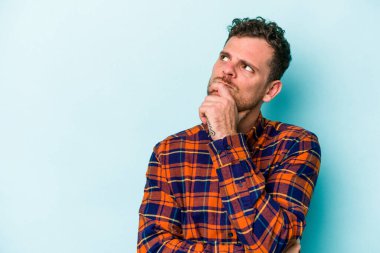 Mavi arka planda izole edilmiş genç beyaz adam şüpheli ve şüpheci bir ifadeyle yana bakıyor..