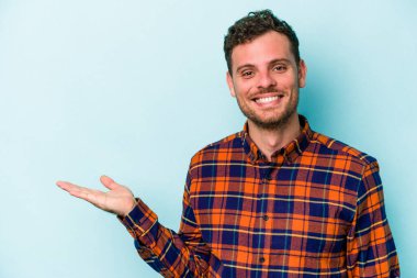 Mavi arka planda izole edilmiş genç beyaz bir adam bir palmiye üzerinde fotokopi alanı ve belinde bir el daha gösteriyor..