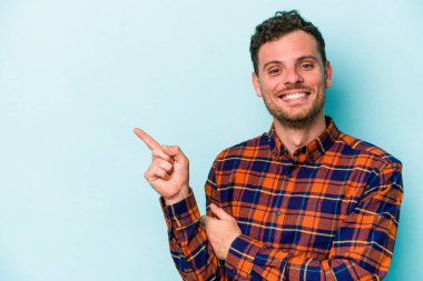 Mavi arka planda izole edilmiş genç beyaz adam işaret parmağıyla işaret ederek gülümsüyor..