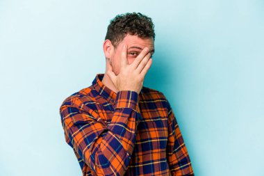 Mavi arka planda izole edilmiş genç beyaz adam kameraya parmaklarıyla göz kırpıyor, yüzünü kapatmaktan utanıyor..