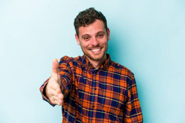 Mavi arka planda izole edilmiş genç beyaz adam selamlama jesti olarak kameraya elini uzatıyor..