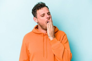 Mavi arka planda izole edilmiş genç beyaz adam esniyor. Ağzı eliyle kapatan yorgun bir hareket sergiliyor..