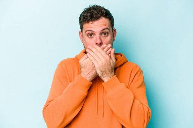 Mavi arka planda izole edilmiş genç beyaz adam elleriyle ağzını kapatırken şok oldu..