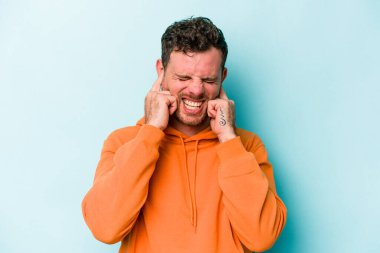 Mavi arka planda izole edilmiş genç beyaz adam kulaklarını elleriyle kapatıyor..