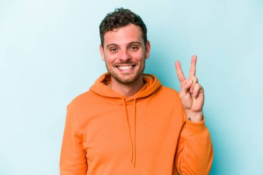 Kafkasyalı genç adam mavi arka planda izole edilmiş neşeli ve kaygısız parmaklarıyla barış sembolü gösteriyor..