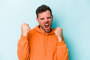 Kafkasyalı genç adam mavi arka planda izole edilmiş kaygısız ve heyecanlı tezahüratlar yapıyor. Zafer konsepti.