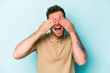 Mavi arka planda soyutlanmış genç beyaz bir adam elleriyle gözlerini kapatıyor, geniş bir gülümsemeyle bir sürpriz bekliyor..