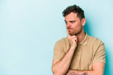 Mavi arka planda izole edilmiş genç beyaz adam şüpheli ve şüpheci bir ifadeyle yana bakıyor..