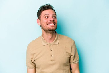 Kafkasyalı genç adam mavi arka planda izole edilmiş rahat ve mutlu bir şekilde gülüyor, boynunda dişlerini gösteriyor..