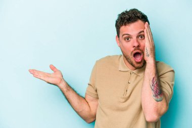 Mavi arka planda izole edilmiş genç beyaz adam, avuç içi fotokopi alanını elinde tutmaktan etkilenmiş..