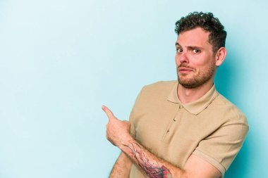 Mavi arka planda izole edilmiş genç beyaz adam gülümsüyor ve boş alanda bir şeyler gösteriyor..