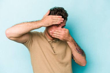 Mavi arka planda izole edilmiş genç beyaz adam kameraya parmaklarıyla göz kırpıyor, yüzünü kapatmaktan utanıyor..