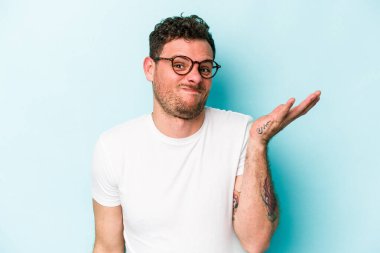 Kafkasyalı genç adam mavi arka planda izole edilmiş sorgulama jestinde şüphe ediyor ve omuz silkiyor..