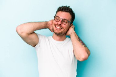 Kafkasyalı genç adam mavi arka planda izole edilmiş. Kendine güveniyor, elleri başının arkasında..