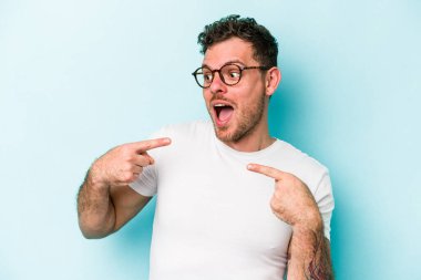 Mavi arka planda izole edilmiş genç beyaz adam parmağını göstererek şaşırdı, geniş bir gülümsemeyle..