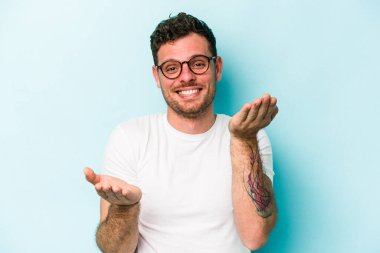 Mavi arka planda izole genç beyaz adam kollarıyla ölçek yapar, mutlu ve kendinden emin hisseder..