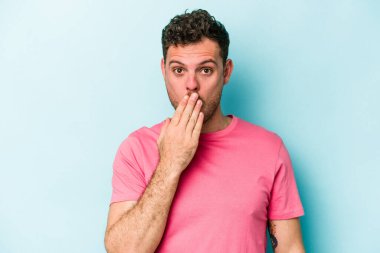 Mavi arka planda izole edilmiş genç beyaz adam şok olmuş, ağzı ellerle kaplanmış, yeni bir şey keşfetmeye hevesli..