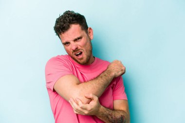 Kafkasyalı genç adam mavi arka planda izole edilmiş dirsek masajı yapıyor, kötü bir hareketin ardından acı çekiyor..