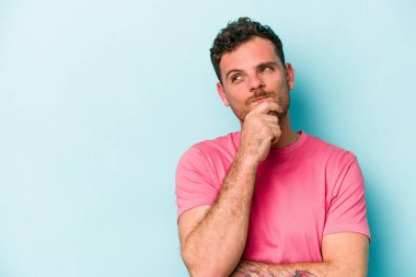 Mavi arka planda izole edilmiş genç beyaz adam şüpheli ve şüpheci bir ifadeyle yana bakıyor..