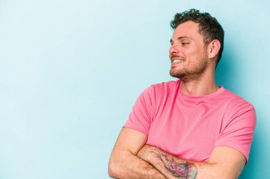 Mavi arka planda izole edilmiş genç beyaz adam kollarını kavuşturarak gülümsüyor..