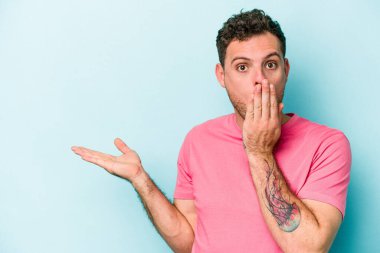 Mavi arka planda izole edilmiş genç beyaz adam, avuç içi fotokopi alanını elinde tutmaktan etkilenmiş..