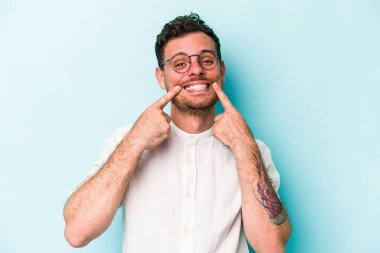 Mavi arka planda izole edilmiş genç beyaz adam gülümsüyor, parmaklarını ağıza doğrultuyor..