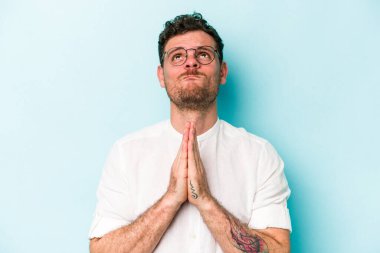 Mavi arka planda izole edilmiş genç beyaz bir adam el ele tutuşup, ağzının kenarında dua ediyor, kendinden emin..