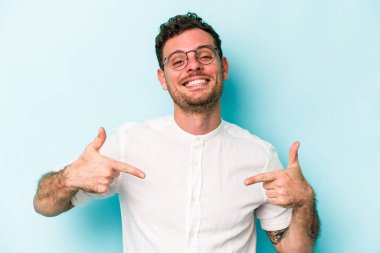 Mavi arka planda izole edilmiş genç beyaz adam bir gömlek fotokopi alanını işaret ediyor, gururlu ve kendinden emin.