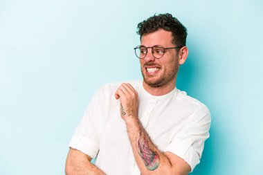 Kafkasyalı genç adam mavi arka planda izole, kafası karışmış, şüpheli ve güvensiz hissediyor..