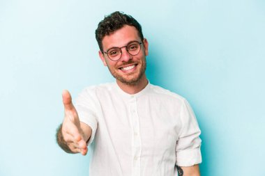 Mavi arka planda izole edilmiş genç beyaz adam selamlama jesti olarak kameraya elini uzatıyor..